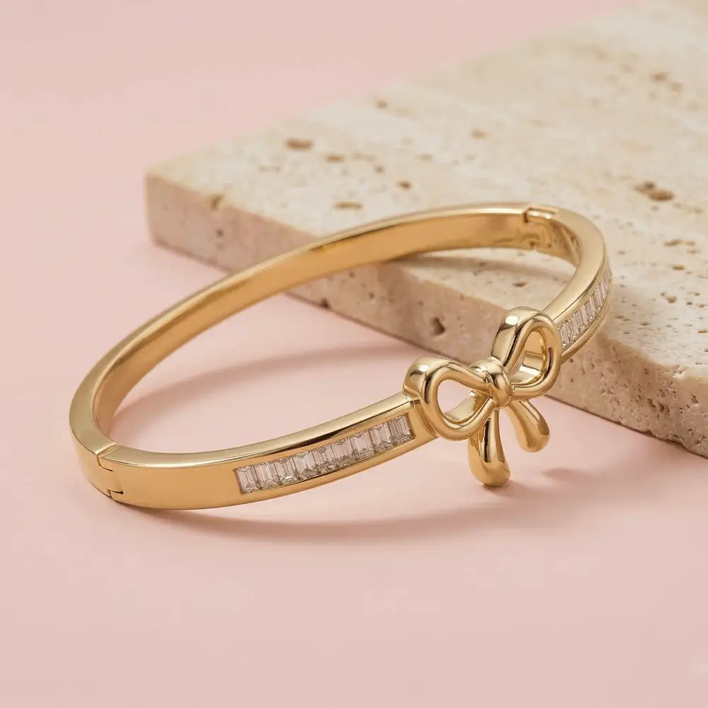 Close-up of a gold bracelet with a polished bow detail and baguette-cut crystal inlays on each side, displayed on a soft pink background with a stone prop.