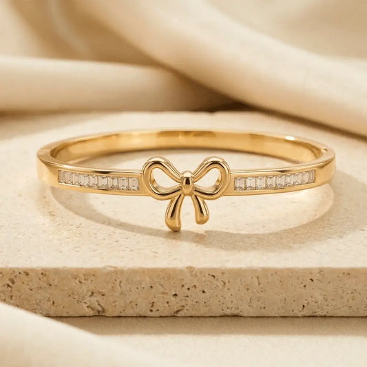 Gold bow bracelet with symmetrical crystal accents on both sides, placed on a textured stone surface with neutral fabric backdrop.