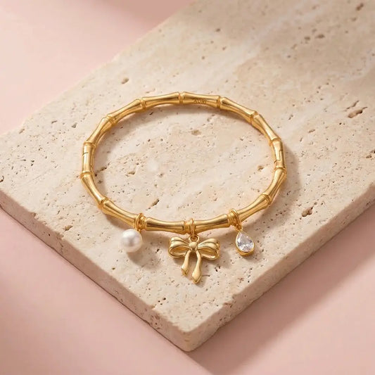 Gold charm bracelet with pearl, bow, and crystal pendant on pastel pink background, anti-tarnish kada bracelet for women