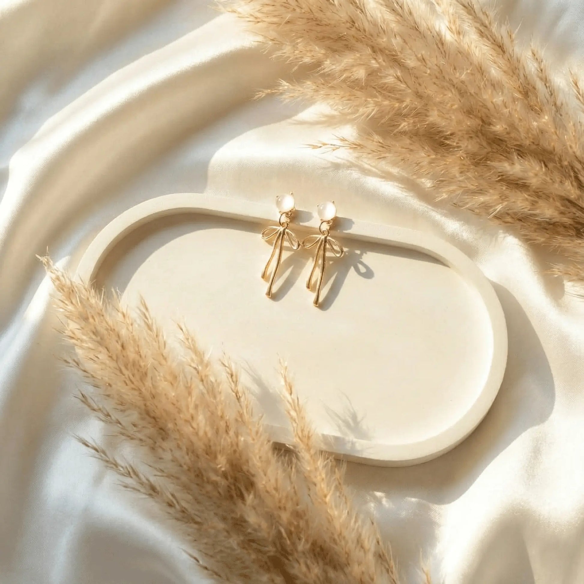 Gold ribbon drop earrings with pearl studs displayed on neutral tray with pampas grass