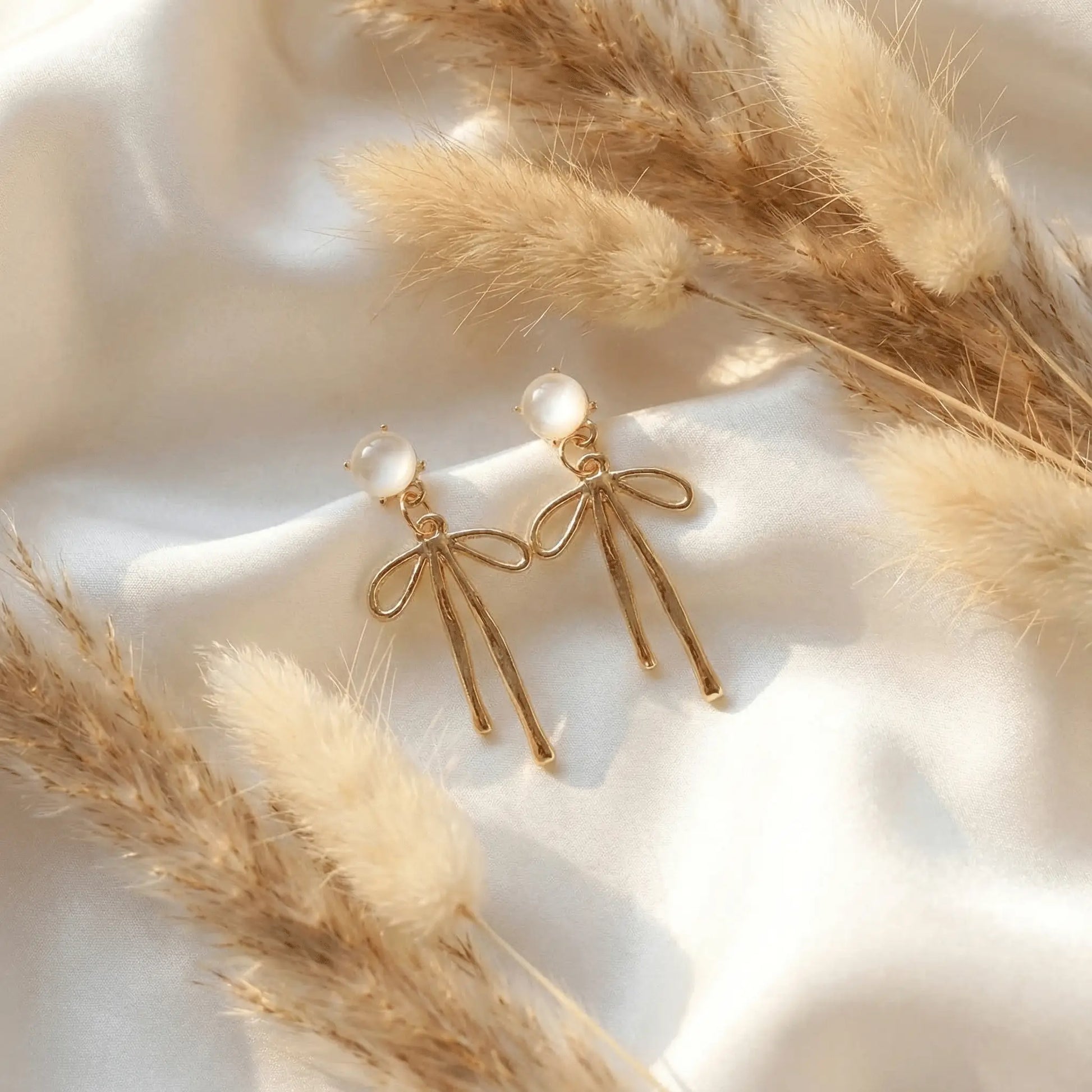 Gold ribbon drop earrings with pearl stud on white satin background, minimal jewellery flat lay