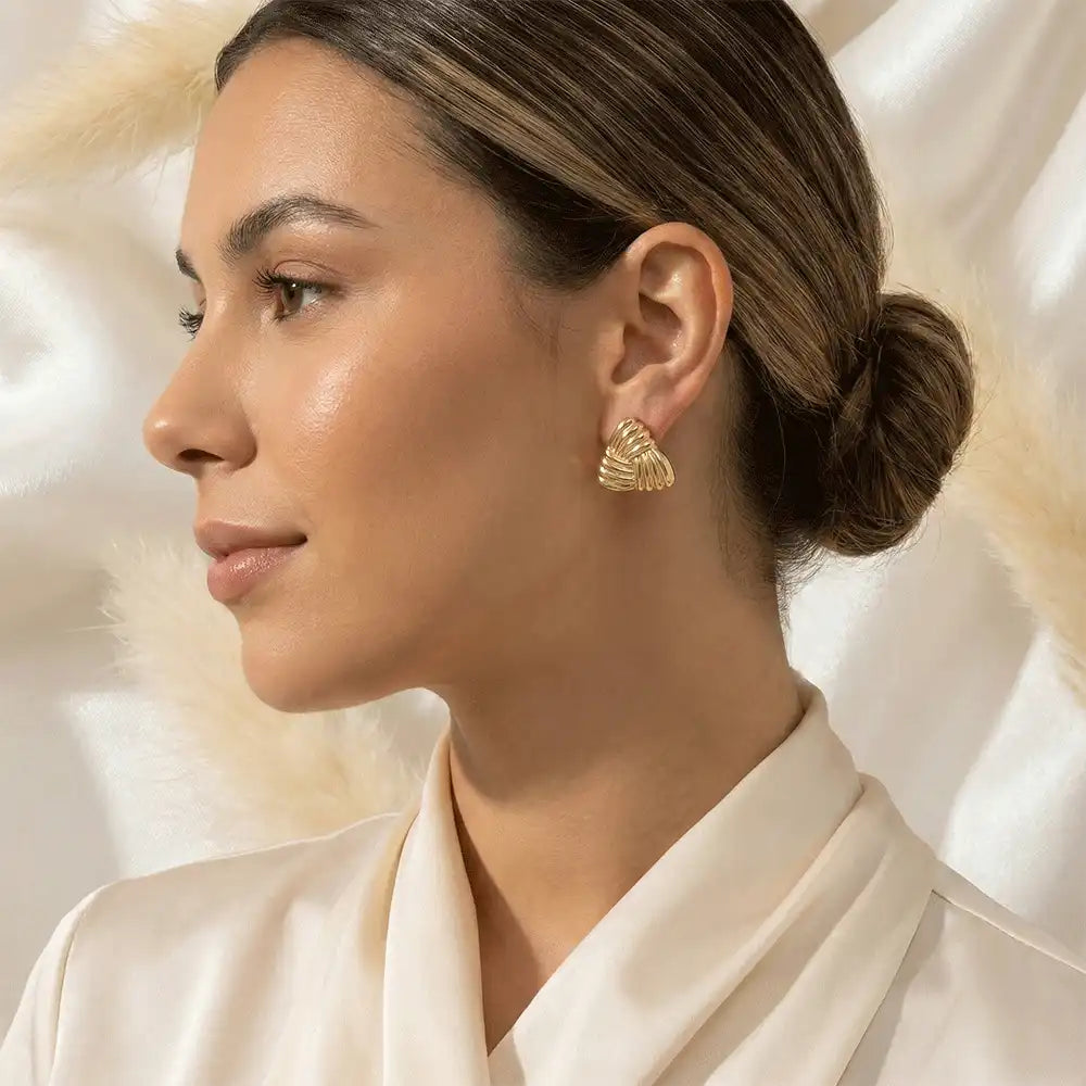 Model wearing gold triangle stud earrings with a textured finish, styled with a low bun hairstyle and soft neutral makeup.