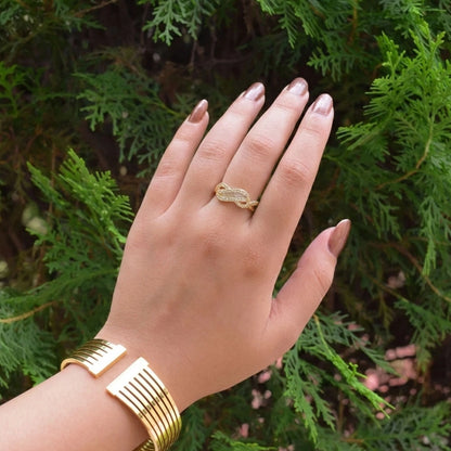 Gold anti-tarnish knot ring worn on hand with minimalist gold bracelet, photographed outdoors against lush green foliage.