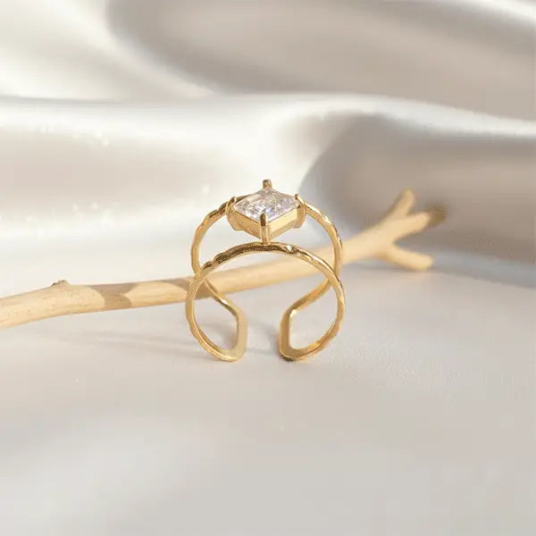 Gold open ring with a raised square-cut crystal centerpiece displayed on soft satin fabric beside a wooden twig.