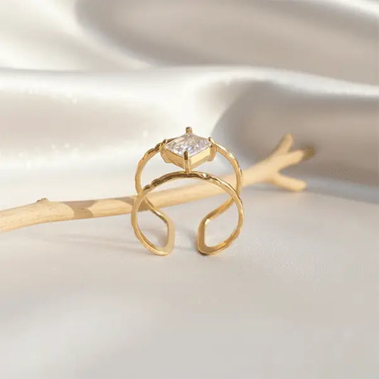 Gold open ring with a raised square-cut crystal centerpiece displayed on soft satin fabric beside a wooden twig.