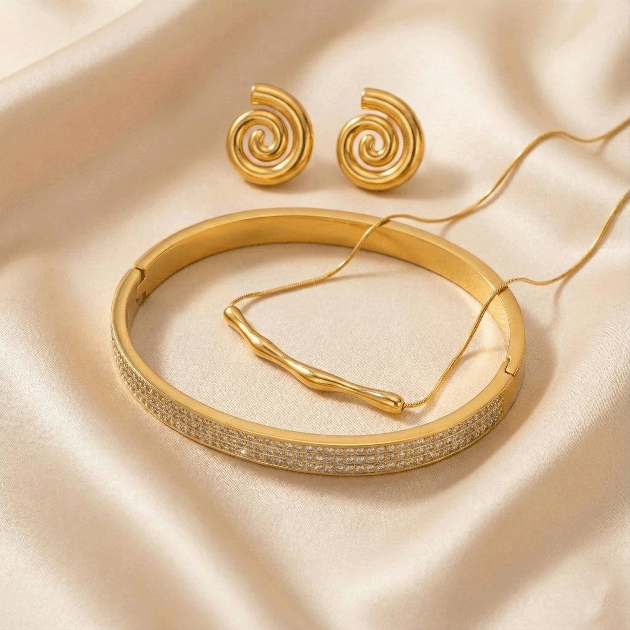 Bold gold jewelry set including spiral stud earrings, a textured gold bangle with crystal detailing, and a sleek curved bar pendant necklace displayed on soft ivory fabric.