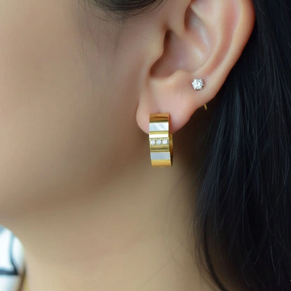 Close-up of gold-toned anti-tarnish hoop earrings with white enamel and crystal detailing worn on ear.