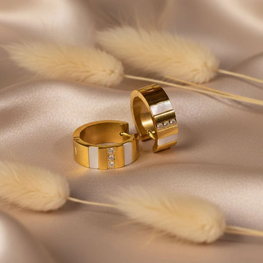 Pair of gold anti-tarnish hoop earrings with white enamel accents placed on satin fabric with pampas grass.