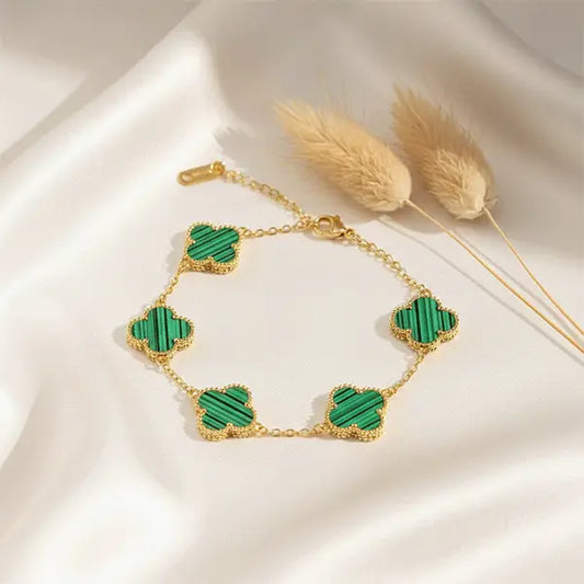 Green clover charm bracelet with gold chain and five malachite-pattern motifs placed on white satin fabric with dried grass props.