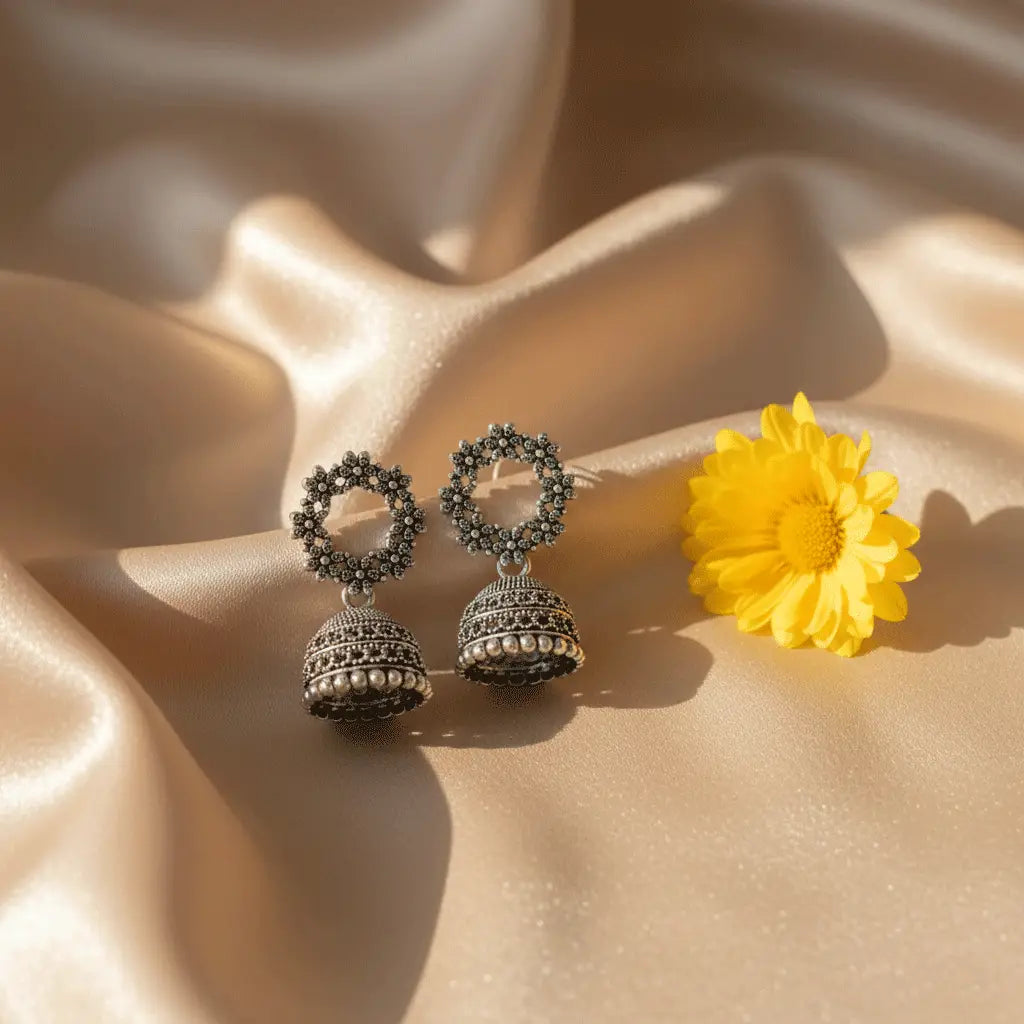 Oxidised silver Gulzar Jhumka earrings placed on beige satin fabric with a yellow flower accent in soft sunlight.”