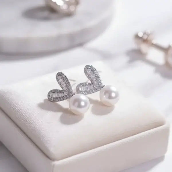 Silver heart shaped stud earrings with baguette-cut stones and round white pearls displayed on a soft jewellery box.