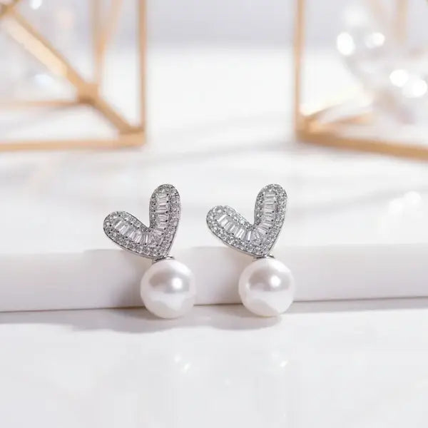 Silver heart-shaped stud earrings with sparkling baguette stones and a dangling white pearl.