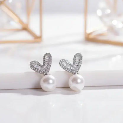 Silver heart-shaped stud earrings with sparkling baguette stones and a dangling white pearl.