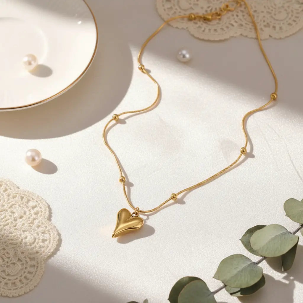 Elegant gold heart pendant necklace with small beads placed on a white surface with pearls and lace decor.