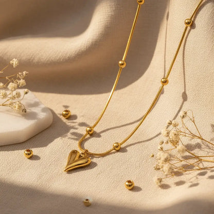 Gold heart pendant necklace with beaded chain displayed on a beige fabric background with soft natural light.