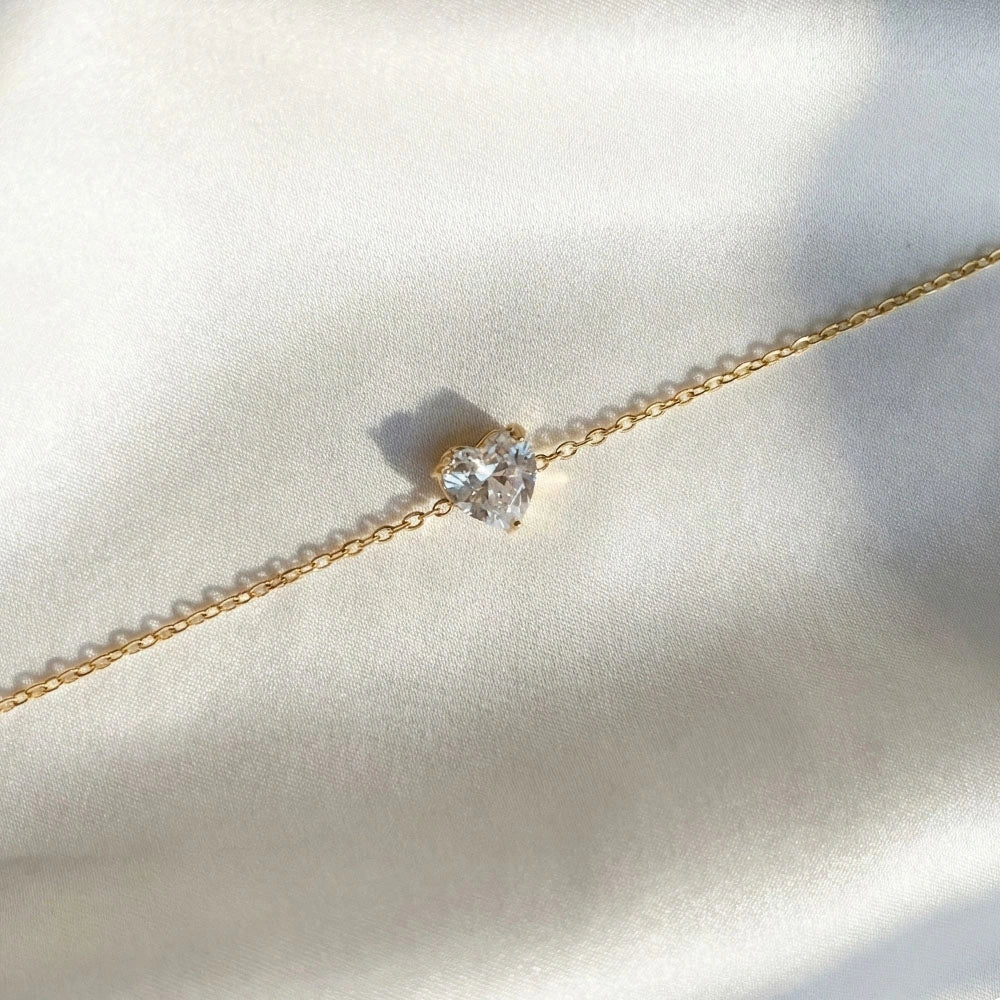 Close-up of gold anti-tarnish bracelet with heart-shaped stone charm laid flat on white satin fabric.