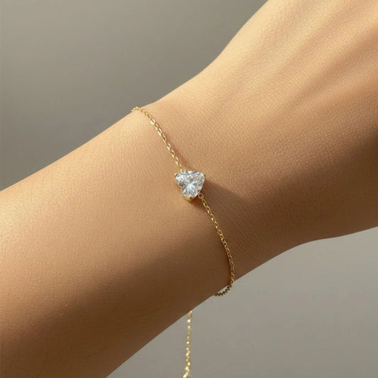 Heart stone charm bracelet worn on wrist, featuring a delicate gold chain and sparkling heart-shaped crystal.