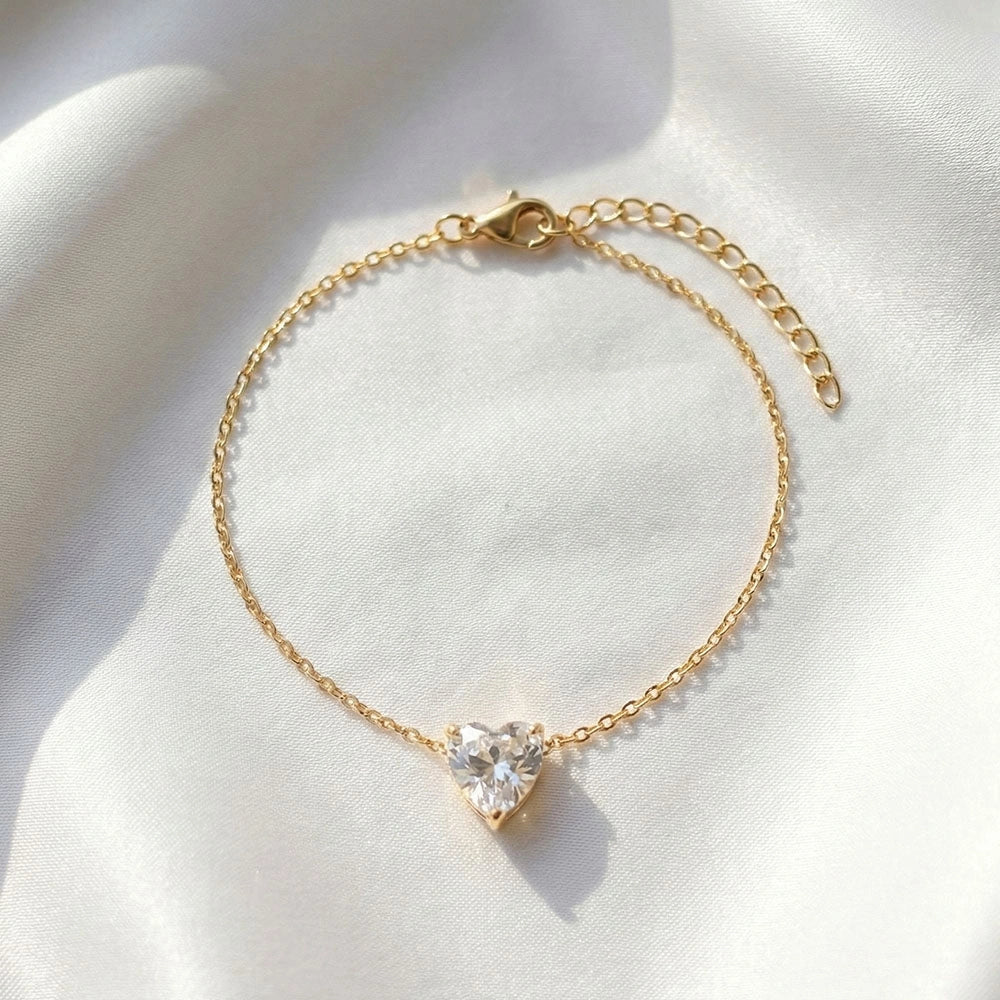 Gold anti-tarnish bracelet with a heart-shaped clear stone charm displayed on soft white fabric background.
