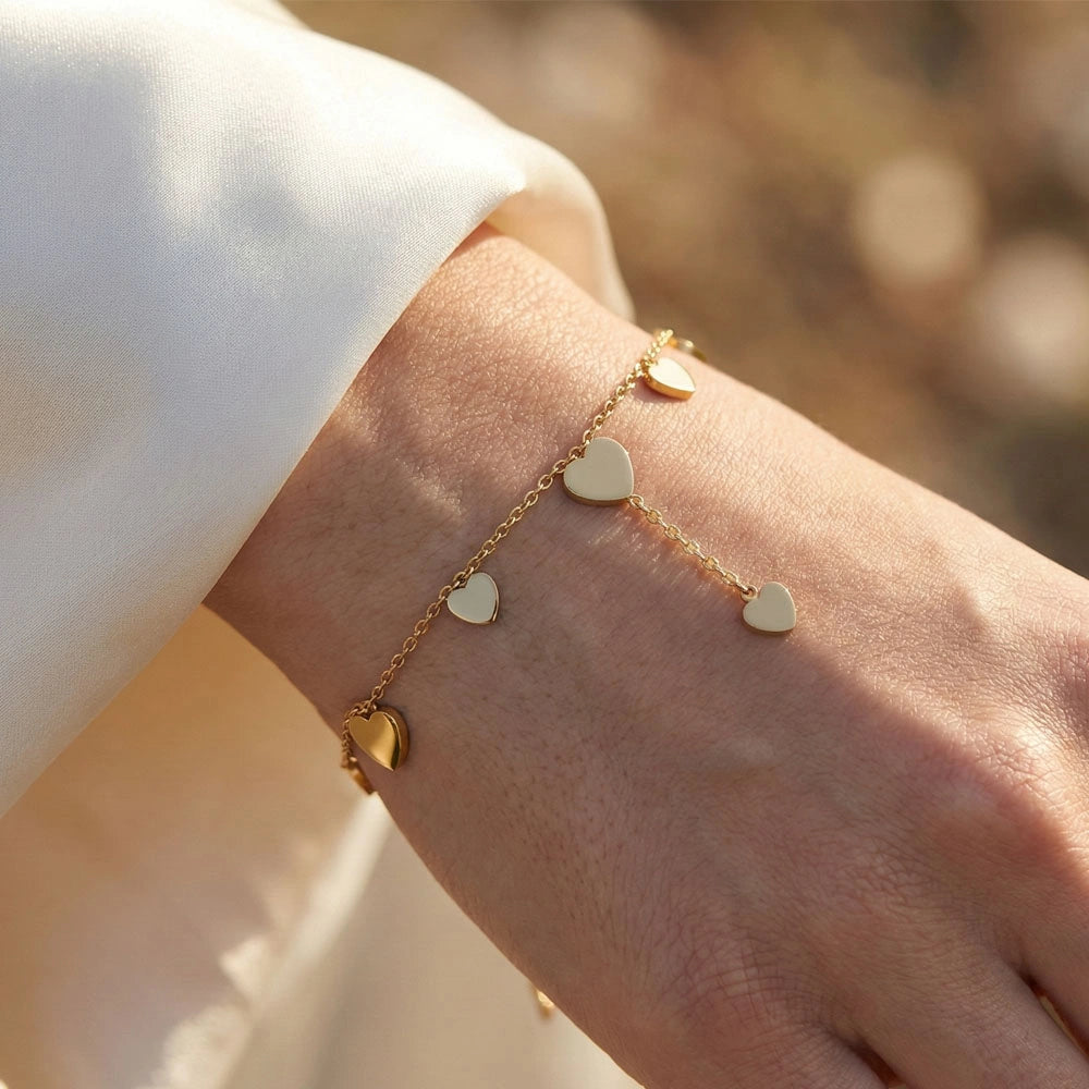Gold heart trail bracelet worn on a wrist, featuring delicate dangling heart charms on a fine chain.