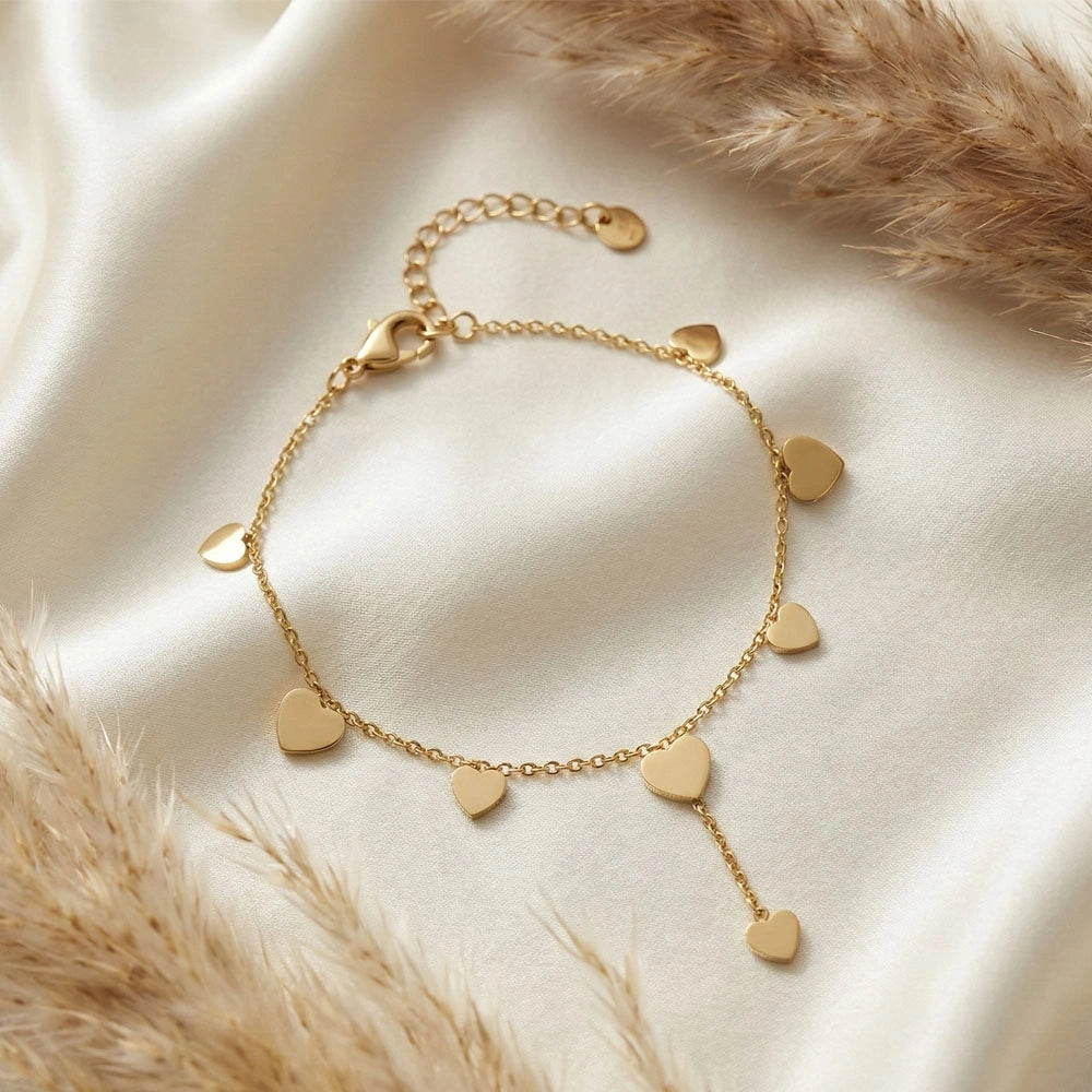 Gold anti-tarnish heart trail bracelet with multiple heart charms arranged on soft ivory fabric with dried florals.