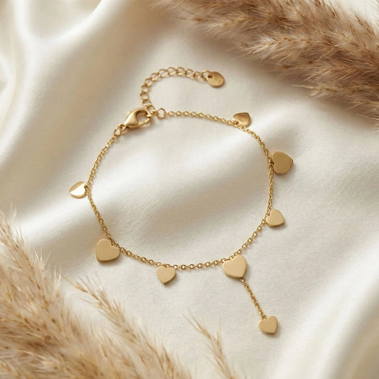 Gold anti-tarnish heart trail bracelet with multiple heart charms arranged on soft ivory fabric with dried florals.