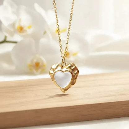 Gold heart-shaped pendant with a white glossy center, hanging on a delicate chain and displayed against soft white orchid flowers on a wooden surface.