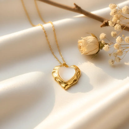 Gold heart-shaped pendant with a white glossy center placed on soft cream satin fabric, styled with a dried rose and delicate baby’s breath flowers in warm natural light.