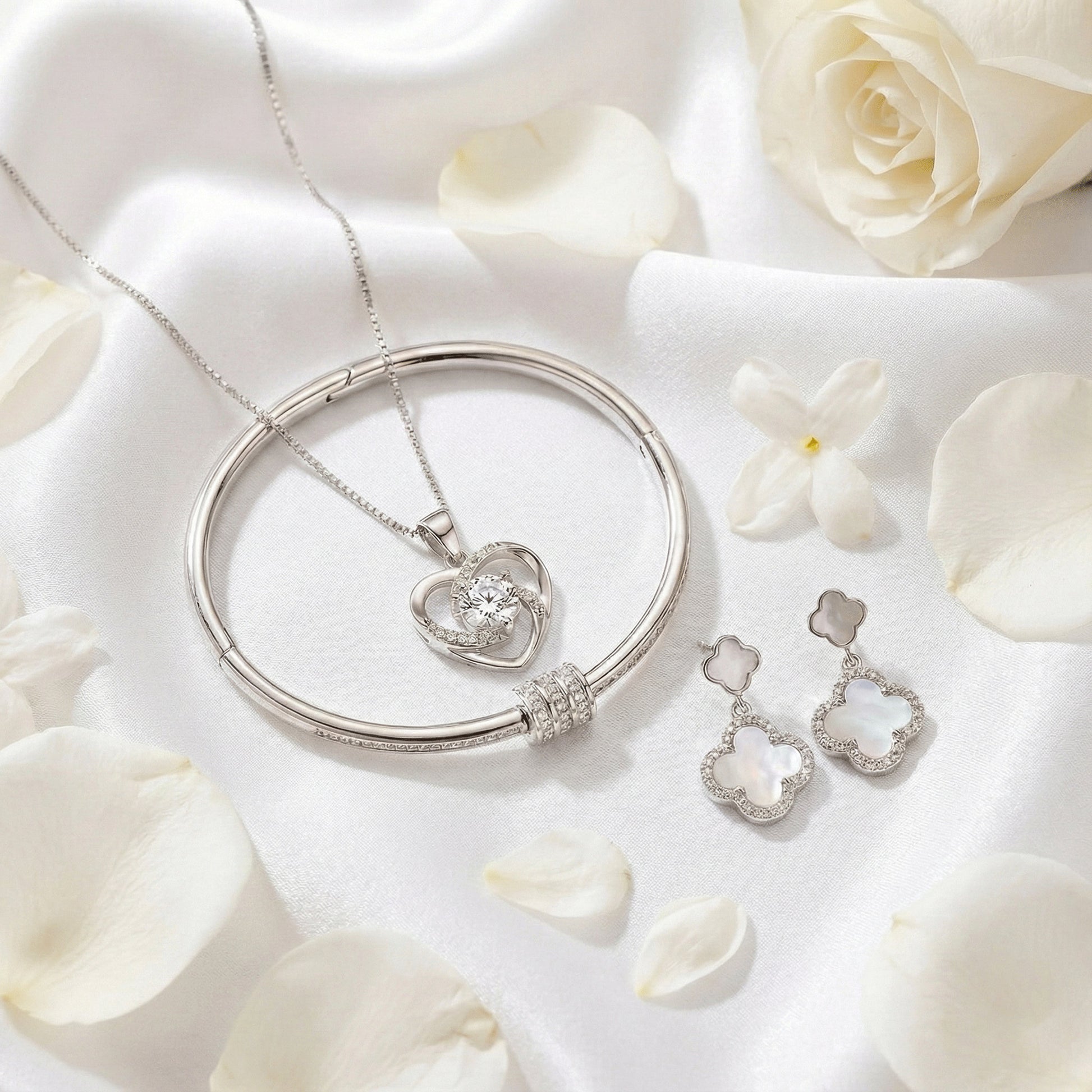 Silver-toned jewelry set featuring a heart-shaped crystal pendant necklace, matching bangle, and clover-inspired drop earrings arranged on white satin with rose petals.