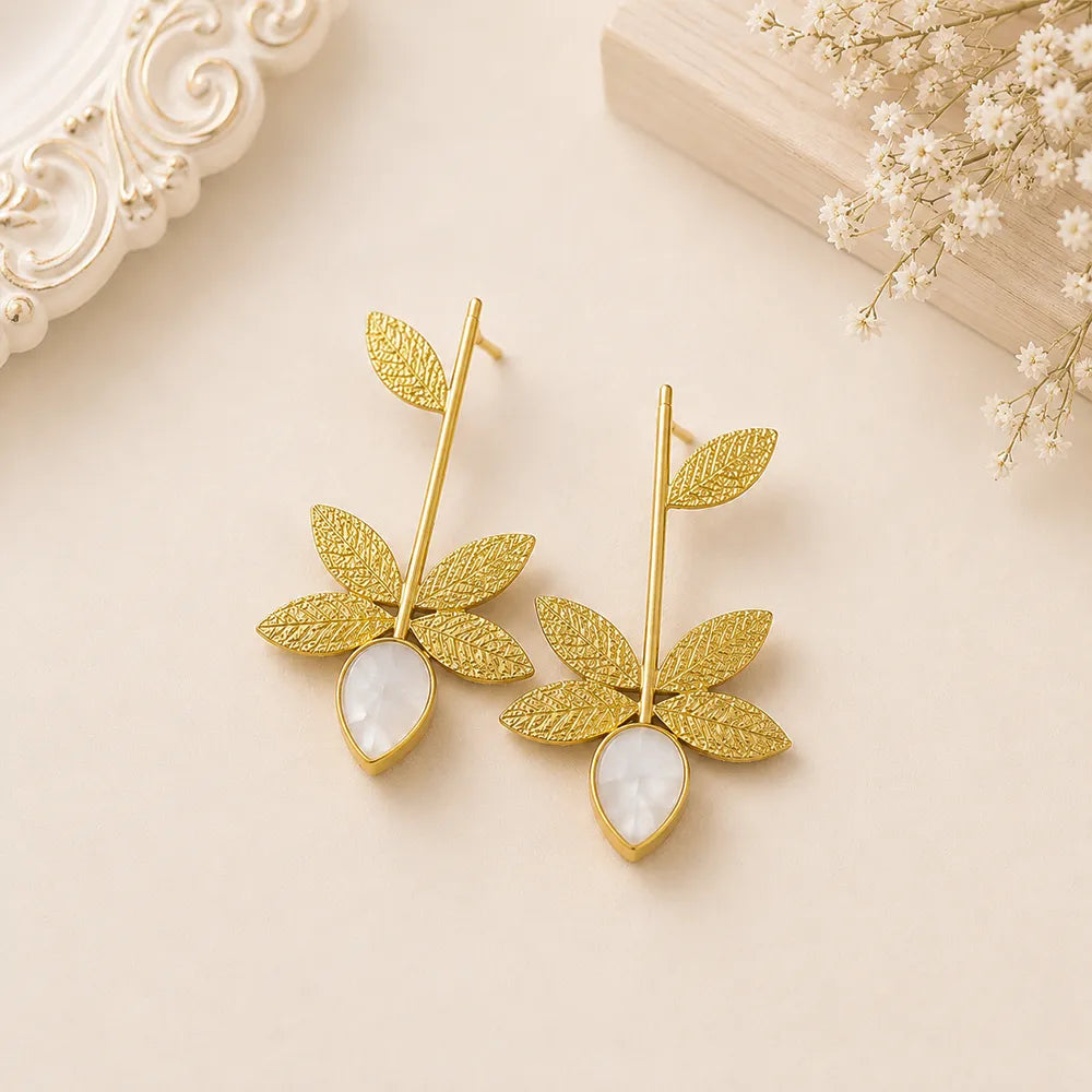 Pair of Ivory Leaf anti tarnish brass earrings with intricate gold-toned leaf design and white teardrop stones on a beige background.