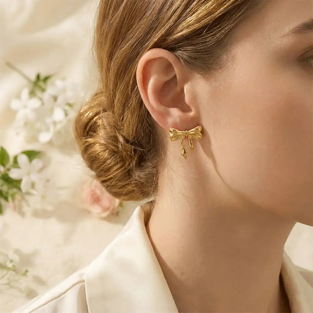 Model wearing gold knot bow stud earrings, styled with a low bun hairstyle and floral background for an elegant, feminine look.