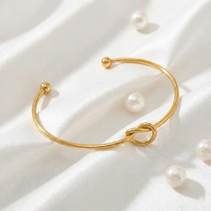 Gold knot bangle bracelet placed on white satin fabric with scattered pearls, highlighting the open cuff design and smooth anti-tarnish finish.