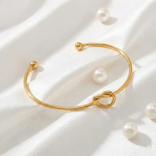 Gold knot bangle bracelet placed on white satin fabric with scattered pearls, highlighting the open cuff design and smooth anti-tarnish finish.