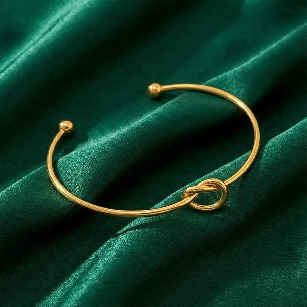 Gold knot bangle bracelet arranged on rich green velvet fabric, showcasing the minimal knot detail and polished gold shine.