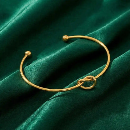 Gold knot bangle bracelet arranged on rich green velvet fabric, showcasing the minimal knot detail and polished gold shine.