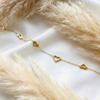 Love Loop Bracelet – Anti-Tarnish, delicate gold heart charm bracelet photographed on white fabric with beige pampas grass accents.