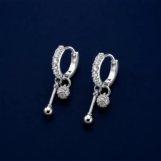 A pair of silver huggie hoop earrings with clear pavé-set stones and dangling ball charms, displayed on a dark navy blue background.