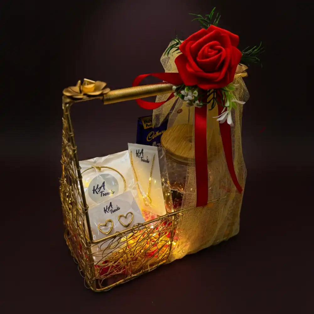 Elegant Valentine gift hamper with anti-tarnish jewellery set, warm fairy lights, chocolates, and a glass jar, showcased in a gold basket with red rose against a dark romantic background.
