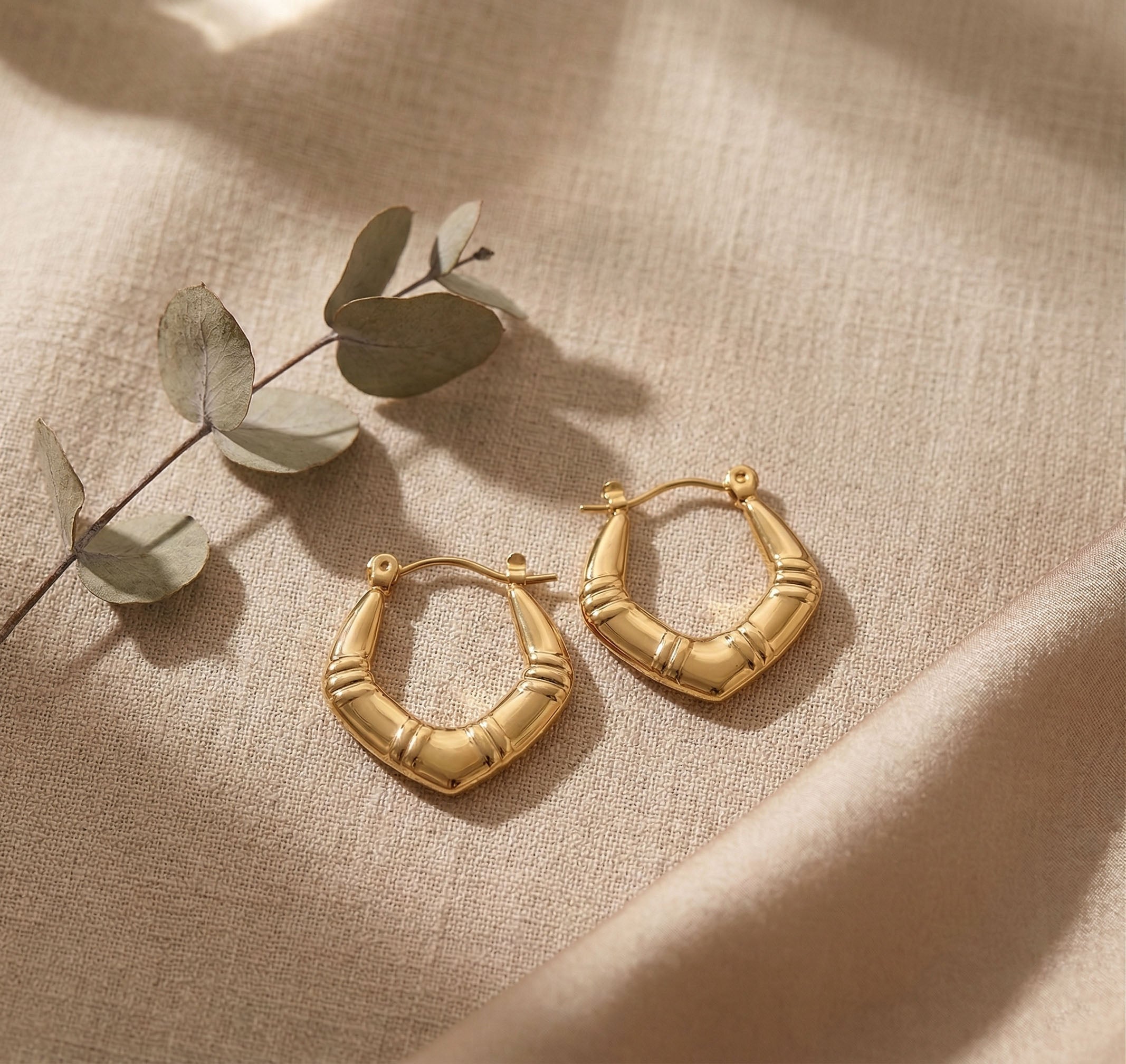 Luxe Arc Hoops on a beige fabric background with a branch of eucalyptus leaves.