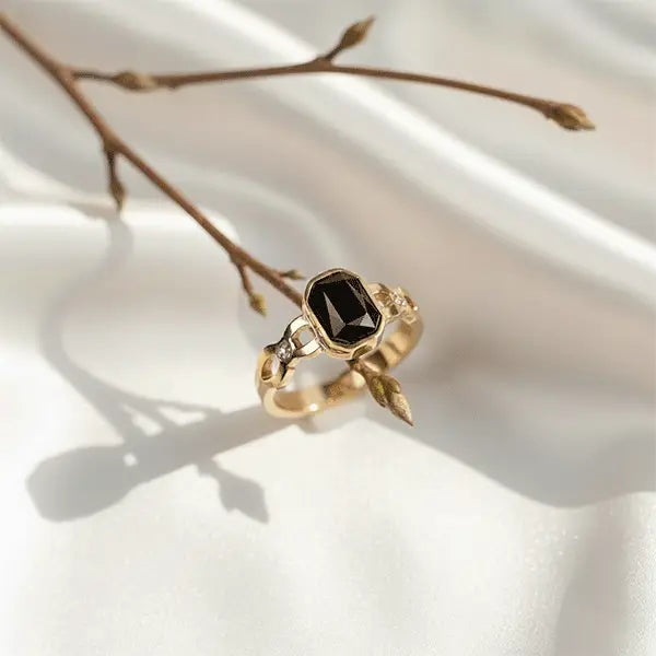 Gold ring with a large black gemstone and leaf-inspired detailing displayed on soft white satin beside a dried branch.