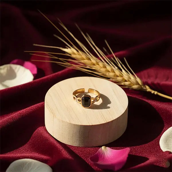 Gold black-stone ring placed on wooden pedestal surrounded by rose petals and wheat on deep red velvet fabric.