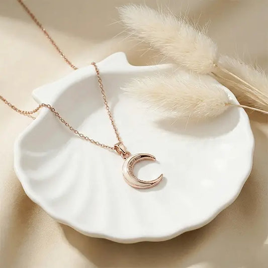 Crescent Moon Pendant necklace in rose-gold finish placed on white ceramic plate with soft sunlight and dried pampas grass