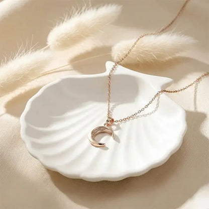 Rose-gold crescent moon pendant necklace styled on white ceramic plate with pampas grass and warm beige background