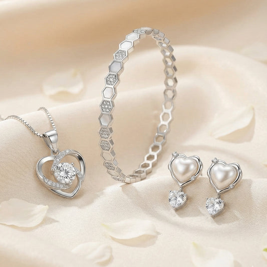 Elegant silver jewelry set with a crystal heart pendant, hexagon-link bracelet, and heart-shaped pearl drop earrings styled on a soft neutral satin background.