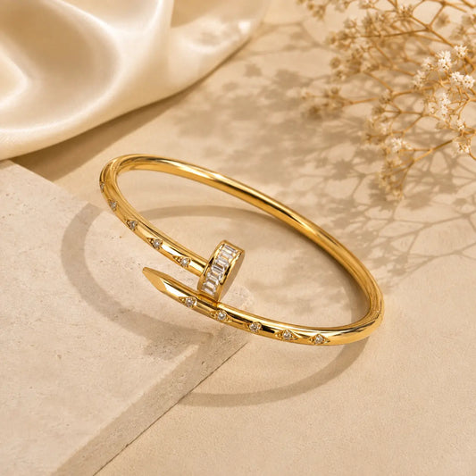A polished Nail Stone Bangle with a central rectangular clear stone cluster and small clear stones along the band, styled on a beige surface with soft shadows and decorative dried flowers.