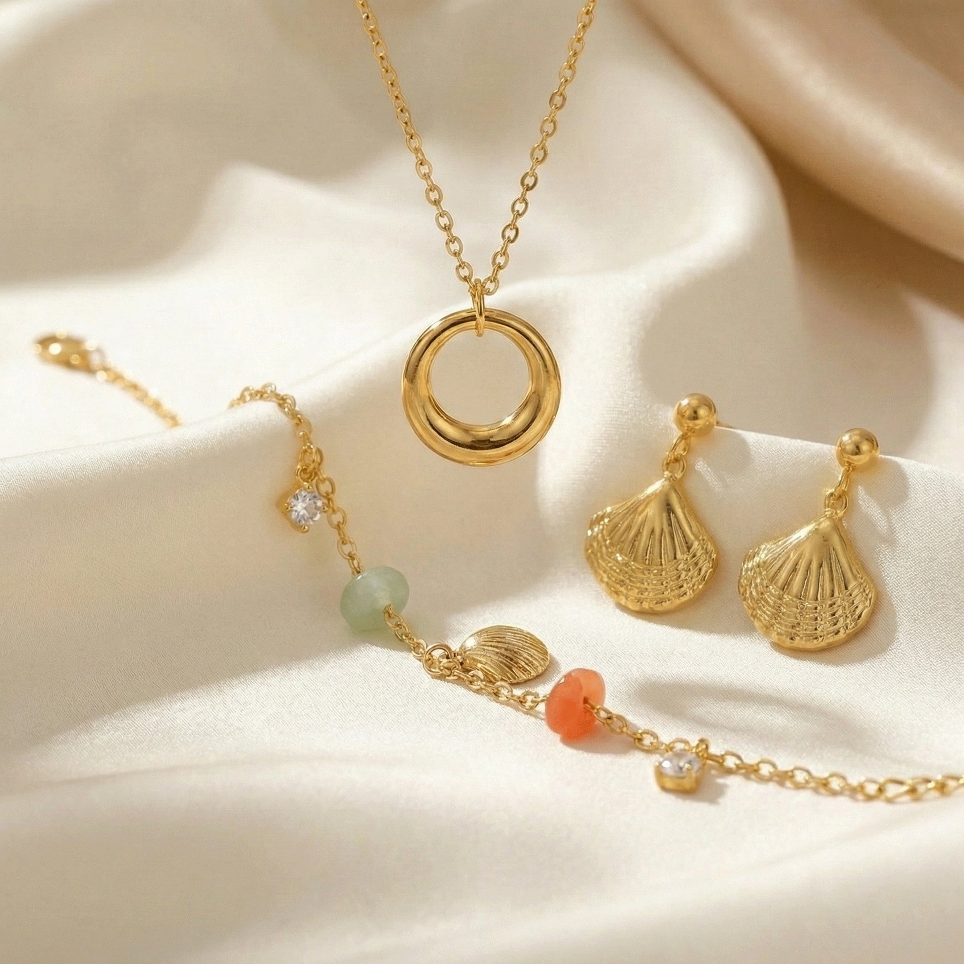 Gold jewelry set featuring a circular pendant necklace, textured shell-shaped drop earrings, and a delicate charm bracelet with pastel beads and crystal accents displayed on soft champagne satin fabric.