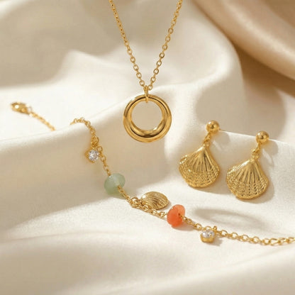 Gold jewelry set featuring a circular pendant necklace, textured shell-shaped drop earrings, and a delicate charm bracelet with pastel beads and crystal accents displayed on soft champagne satin fabric.