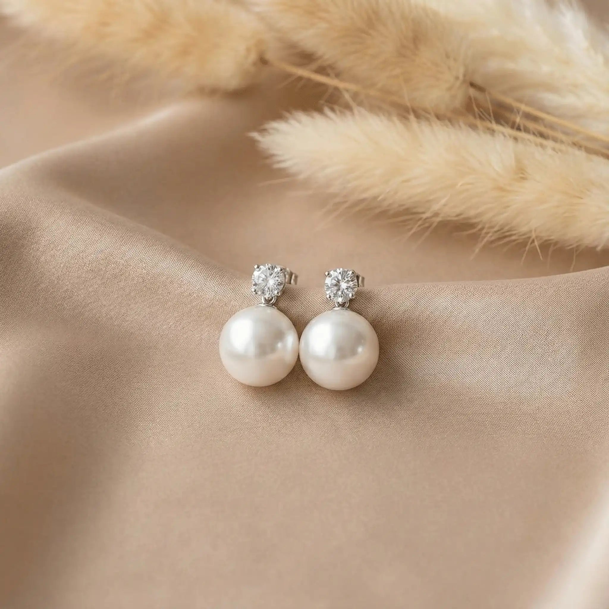 Pearl drop earrings with crystal studs placed on beige satin fabric, minimal luxury jewellery shot