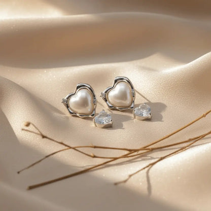 Silver-plated heart-shaped pearl stud earrings with detached cubic zirconia heart drop charms on soft beige satin fabric. Elegant, romantic jewelry.