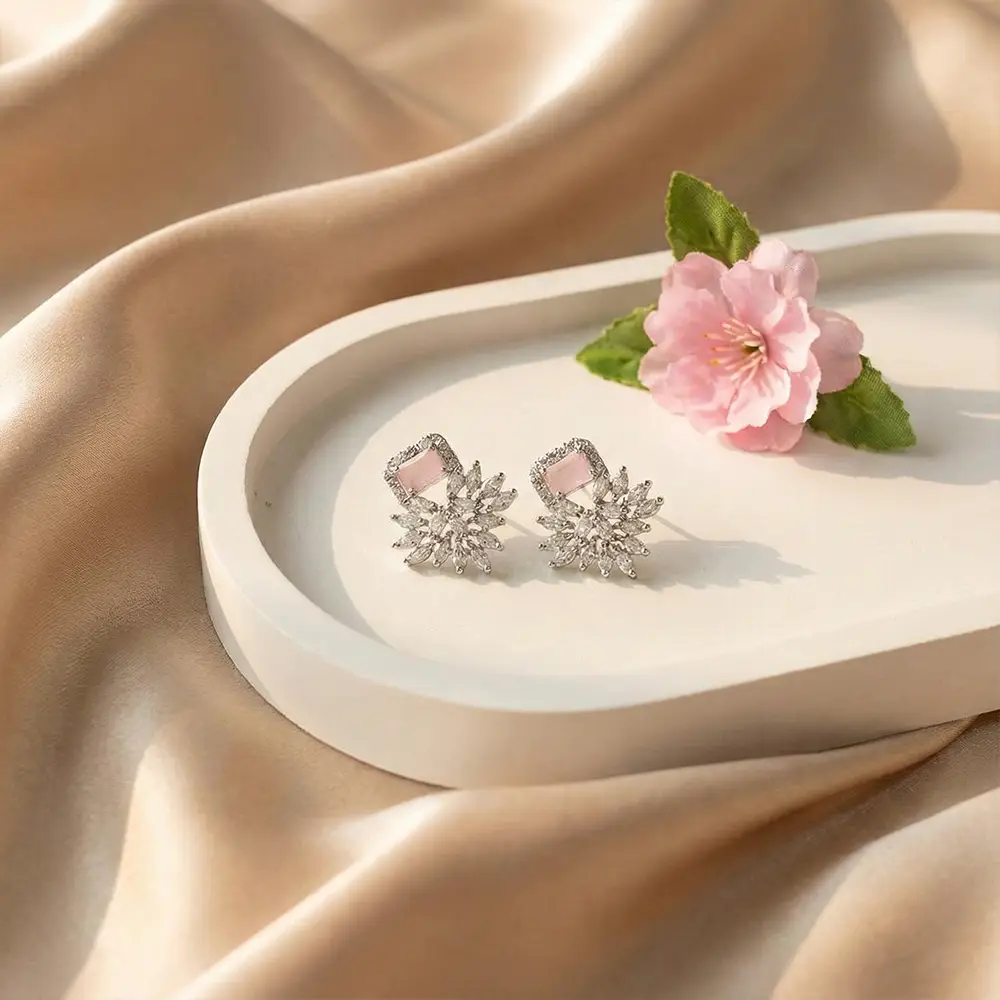 A pair of Pink Petal American diamond stud earrings displayed on white tray with a pink flower decoration, set against a silky gold fabric background.