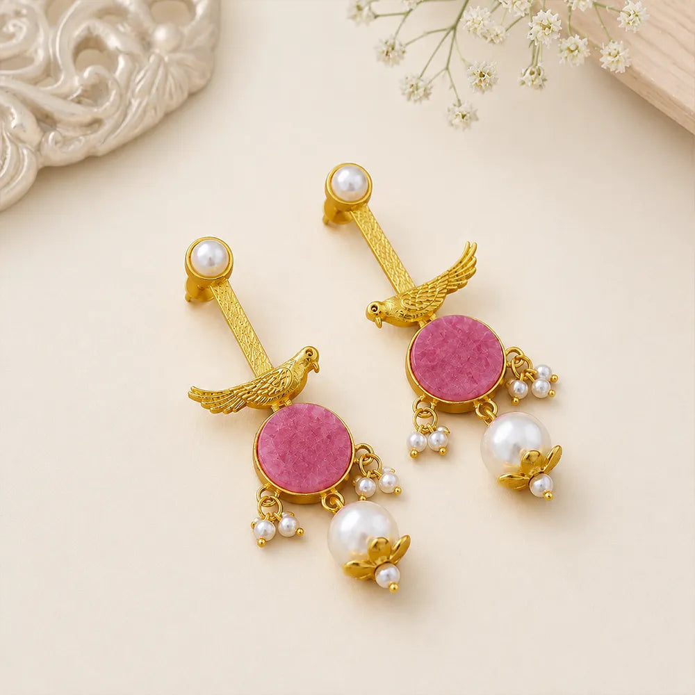 : Pair of Rosy Sparrow anti tarnish brass earrings with pink round stones, gold-toned bird motifs, and pearl danglers on a beige background.